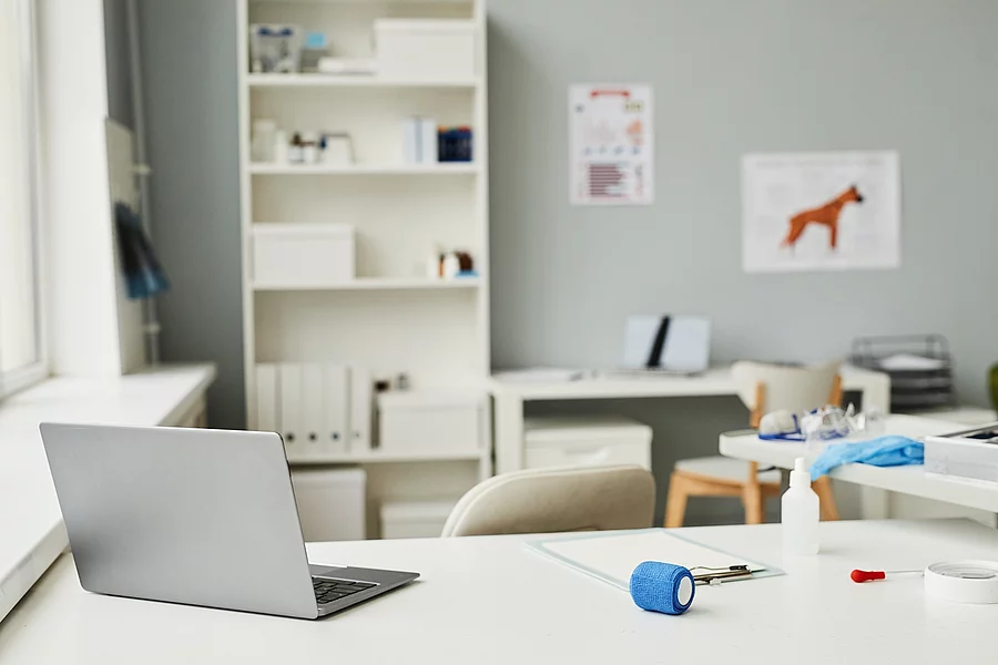 Arbeitsplatz in einer Tierarztpraxis. Im Vordergrund des Bildes steht ein weißer Schreibtisch. Auf dem Tisch ist ein geöffneter Laptop abgestellt und weitere Utensilien - wie eine Bandage und ein Klemmbrett - liegen auf dem Tisch. Im Hintergrund steht eine Bücherregal und medizinische Plakate hängen an der Wand.