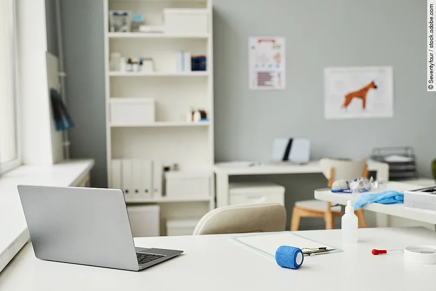 Arbeitsplatz in einer Tierarztpraxis. Im Vordergrund des Bildes steht ein weißer Schreibtisch. Auf dem Tisch ist ein geöffneter Laptop abgestellt und weitere Utensilien - wie eine Bandage und ein Klemmbrett - liegen auf dem Tisch. Im Hintergrund steht eine Bücherregal und medizinische Plakate hängen an der Wand.