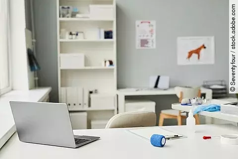 Arbeitsplatz in einer Tierarztpraxis. Im Vordergrund des Bildes steht ein weißer Schreibtisch. Auf dem Tisch ist ein geöffneter Laptop abgestellt und weitere Utensilien - wie eine Bandage und ein Klemmbrett - liegen auf dem Tisch. Im Hintergrund steht eine Bücherregal und medizinische Plakate hängen an der Wand.