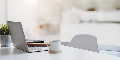 Auf einem weißen Schreibtisch steht ein silberner Laptop aufgeklappt. Neben dem Laptop steht eine Kaffeetasse auf einem Untersetzer und hinter dem Laptop ist eine kleine grüne Zimmerpflanze platziert. An den Tisch ist ein Stuhl geschoben.