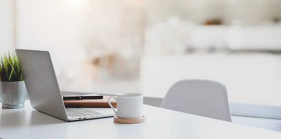 Modern workplace with laptop computer, coffee cup and office sup Auf einem weißen Schreibtisch steht ein silberner Laptop aufgeklappt. Neben dem Laptop steht eine Kaffeetasse auf einem Untersetzer und hinter dem Laptop ist eine kleine grüne Zimmerpflanze platziert. An den Tisch ist ein Stuhl geschoben.
