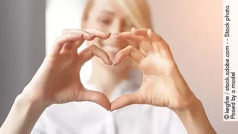 Eine blonde Frau mit weißem Oberteil formt mit ihren beiden Händen ein Herz und lächelt in die Kamera. Eine blonde Frau mit weißem Oberteil formt mit ihren beiden Händen ein Herz und lächelt in die Kamera.
