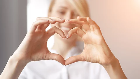 Eine blonde Frau mit weißem Oberteil formt mit ihren beiden Händen ein Herz und lächelt in die Kamera.