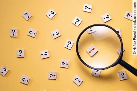 Mit einer Lupe wird ein Holzstein mit einem roten Ausrufezeichen vergrößert dargestellt. Um diesen Stein herum liegen nur Steine mit schwarzen Fragezeichen auf gelbem Hintergrund.