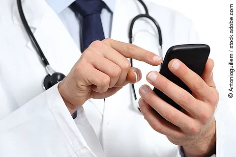 Ein Arzt hält mit der linken Hand ein schwarzes Smartphone. Mit dem Zeigefinger der rechten Hand bedient er das Handy. Er trägt einen weißen Kittel und darunter ein hellblaues Hemd mit dunkelblauer Krawatte. Um seinen Hals hängt ein Stethoskop.