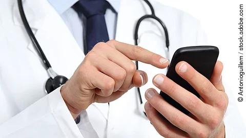 Ein Arzt hält mit der linken Hand ein schwarzes Smartphone. Mit dem Zeigefinger der rechten Hand bedient er das Handy. Er trägt einen weißen Kittel und darunter ein hellblaues Hemd mit dunkelblauer Krawatte. Um seinen Hals hängt ein Stethoskop.