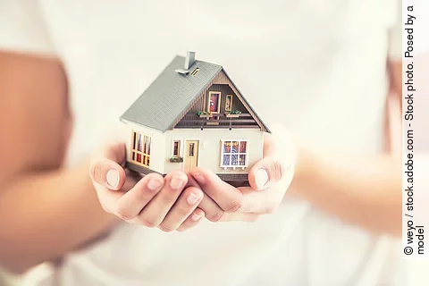 Eine junge Frau hält ein kleines Modellhaus schützend in ihren Händen.