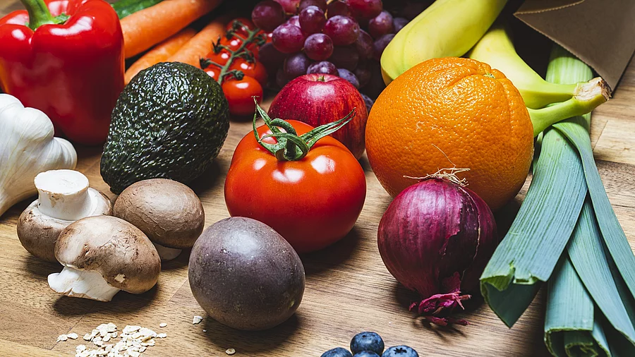 Auf einem Holztisch liegt buntes Gemüse und Obst. Darunter sind braune Chamignons, eine Avocado, rote Paprika und Lauch. 