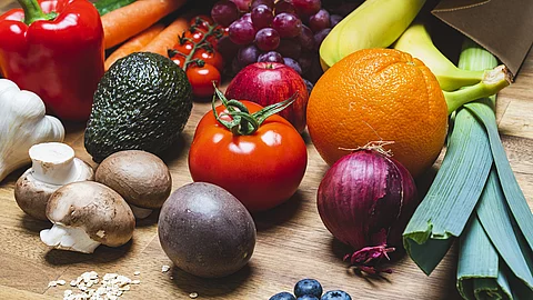 Auf einem Holztisch liegt buntes Gemüse und Obst. Darunter sind braune Chamignons, eine Avocado, rote Paprika und Lauch. 