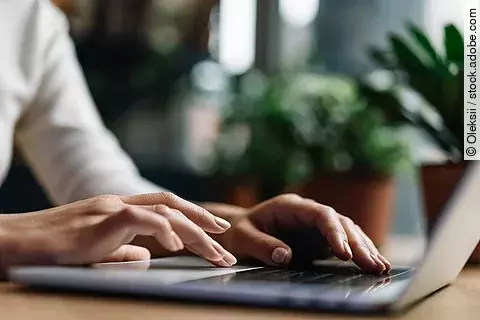 Nahaufnahme von Händen, die einen Laptop benutzen und auf der Tastatur tippen. Der Laptop ist silber und im Hintergrund stehen grüne Pflanzen. 