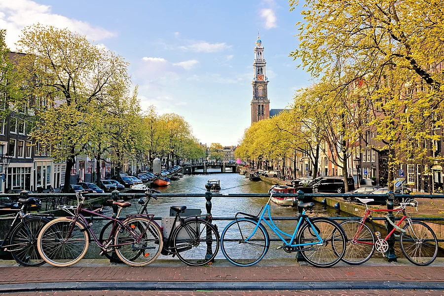Fahrräder lehnen an einer Brücke über die Kanäle von Amsterdam, im Hintergrund eine Kirche. Aufnahme von einer Brücke über einen Fluss in Amsterdam. Seitlich vom Fluss stehen grüne Bäume. Fahrräder lehnen an einer Brücke über die Kanäle von Amsterdam, im Hintergrund eine Kirche. Es herrscht spätes Tageslicht.