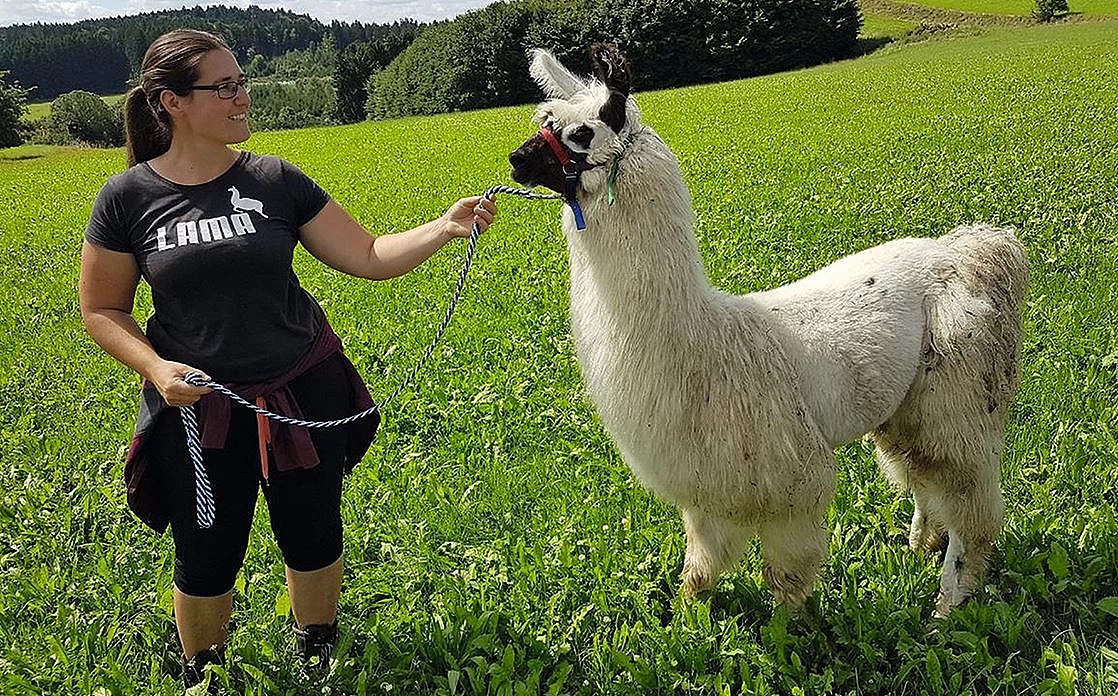 Anja Kaindl steht auf einer grünen Wiese und hält ein weißes Alpaka mit Halfter und Strick.