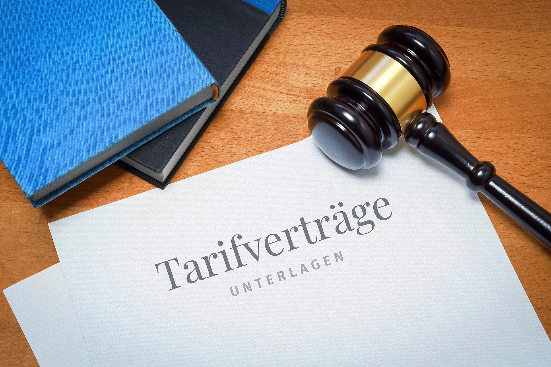 Auf einem Holztisch liegt ein Dokument. Auf der Vorderseite des Dokuments steht "Tarifverträge Unterlagen". Rechts der Tarifverträge leit ein Richterhammer und oberhalb sind die Kanten von einem blauen und einem schwarzen Buch erkennbar. 