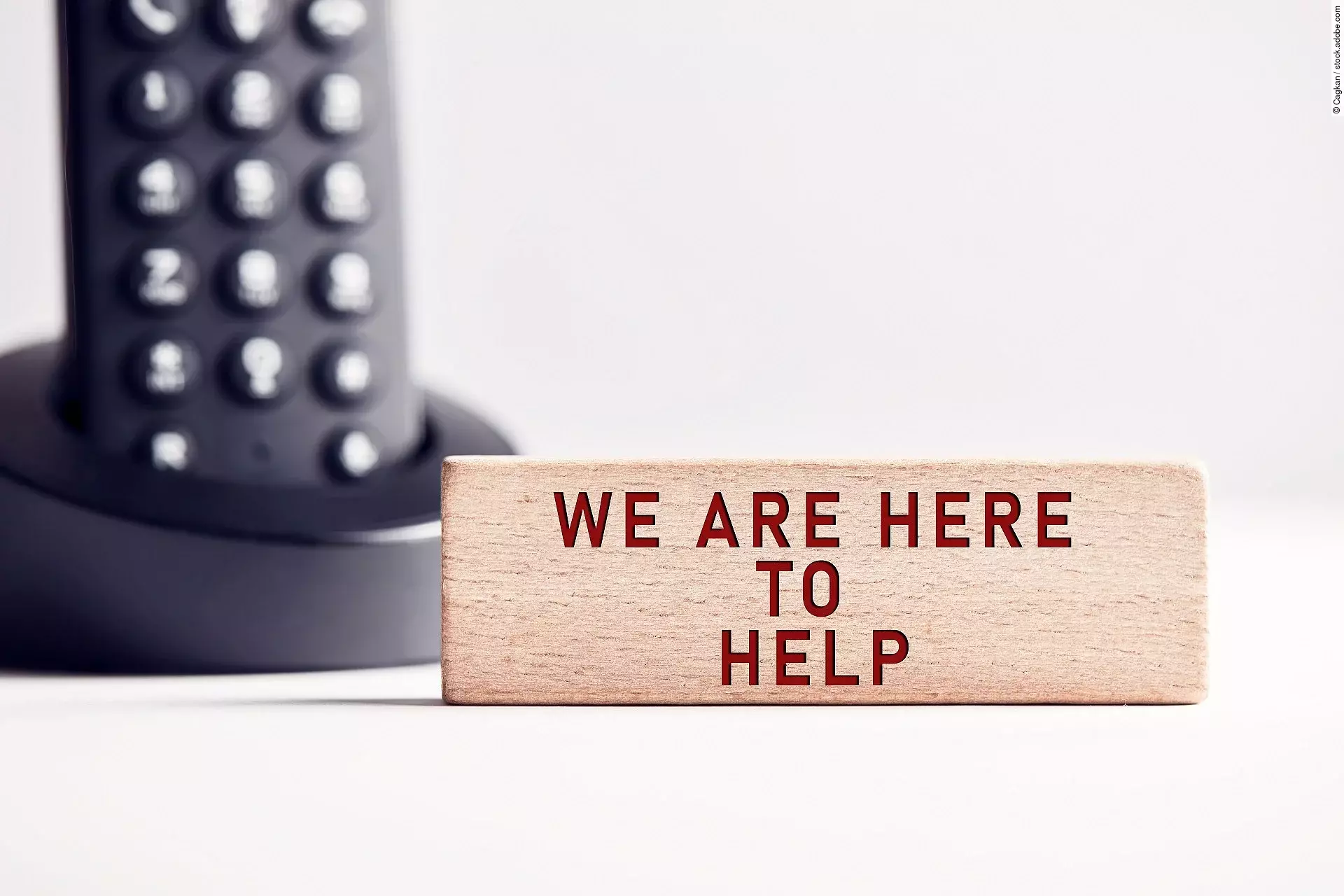 Der Satz "We are here to help" steht in roten Buchstaben auf einem Holzblock. Im Hintergrund steht ein schwarzes Telefon.