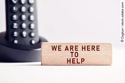 Der Satz "We are here to help" steht in roten Buchstaben auf einem Holzblock. Im Hintergrund steht ein schwarzes Telefon.