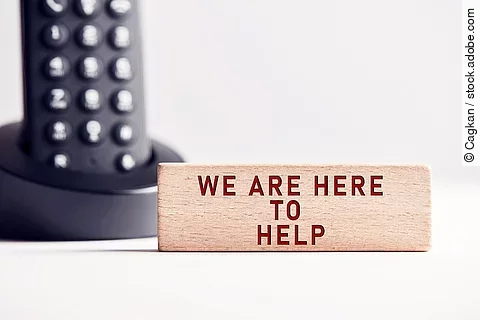 Der Satz "We are here to help" steht in roten Buchstaben auf einem Holzblock. Im Hintergrund steht ein schwarzes Telefon.