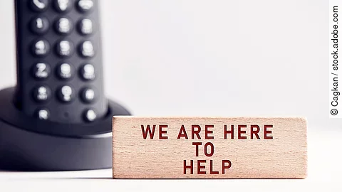Der Satz "We are here to help" steht in roten Buchstaben auf einem Holzblock. Im Hintergrund steht ein schwarzes Telefon.