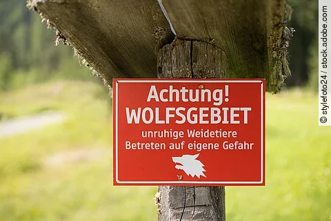 Rotes Warnschild an einem Baum an einer Wiese mit dem Text: Achtung Wolfsgebiet, Betreten auf eigene Gefahr.