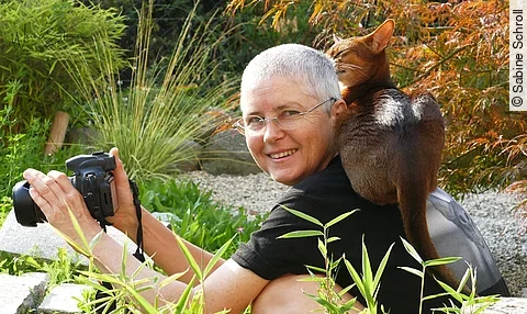Sabine Schroll sitzt mit einer Kamera in der Hand und einer Katze auf der Schulter in der Natur.