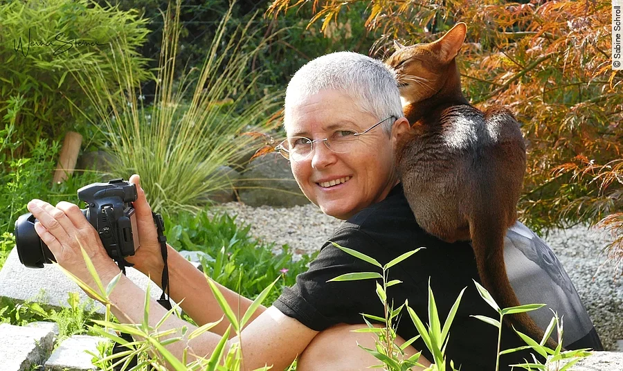 Sabine Schroll sitzt mit einer Kamera in der Hand und einer Katze auf der Schulter in der Natur.