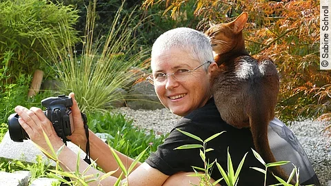 Sabine Schroll sitzt mit einer Kamera in der Hand und einer Katze auf der Schulter in der Natur.