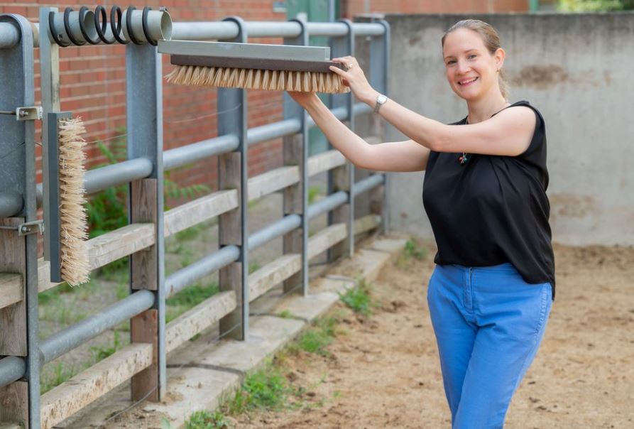 Borbala Foris steht lächelnd in einem Auslauf für Rinder und hält eine Bürste zum Kratzen für die Tiere fest. Dabei sieht sie glücklich aus. 