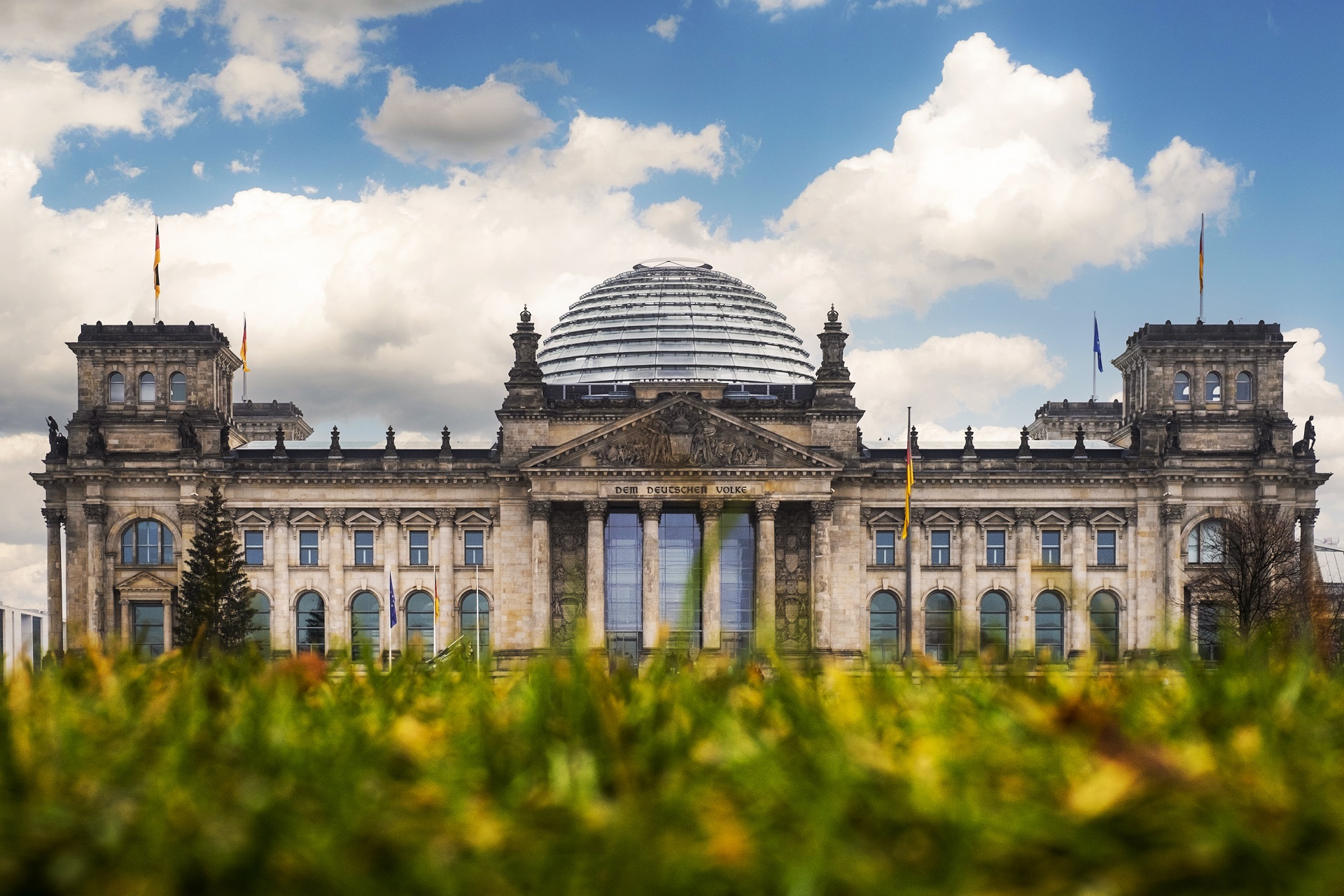 Aufnahme vom Reichstagsgebäude in Berlin. Das Gebäude ist von unten fotografiert und im Hintergrund ist ein blauer Himmel mit Wolken. Im Vordergrund des Bildes ist eine grüne Wiese.