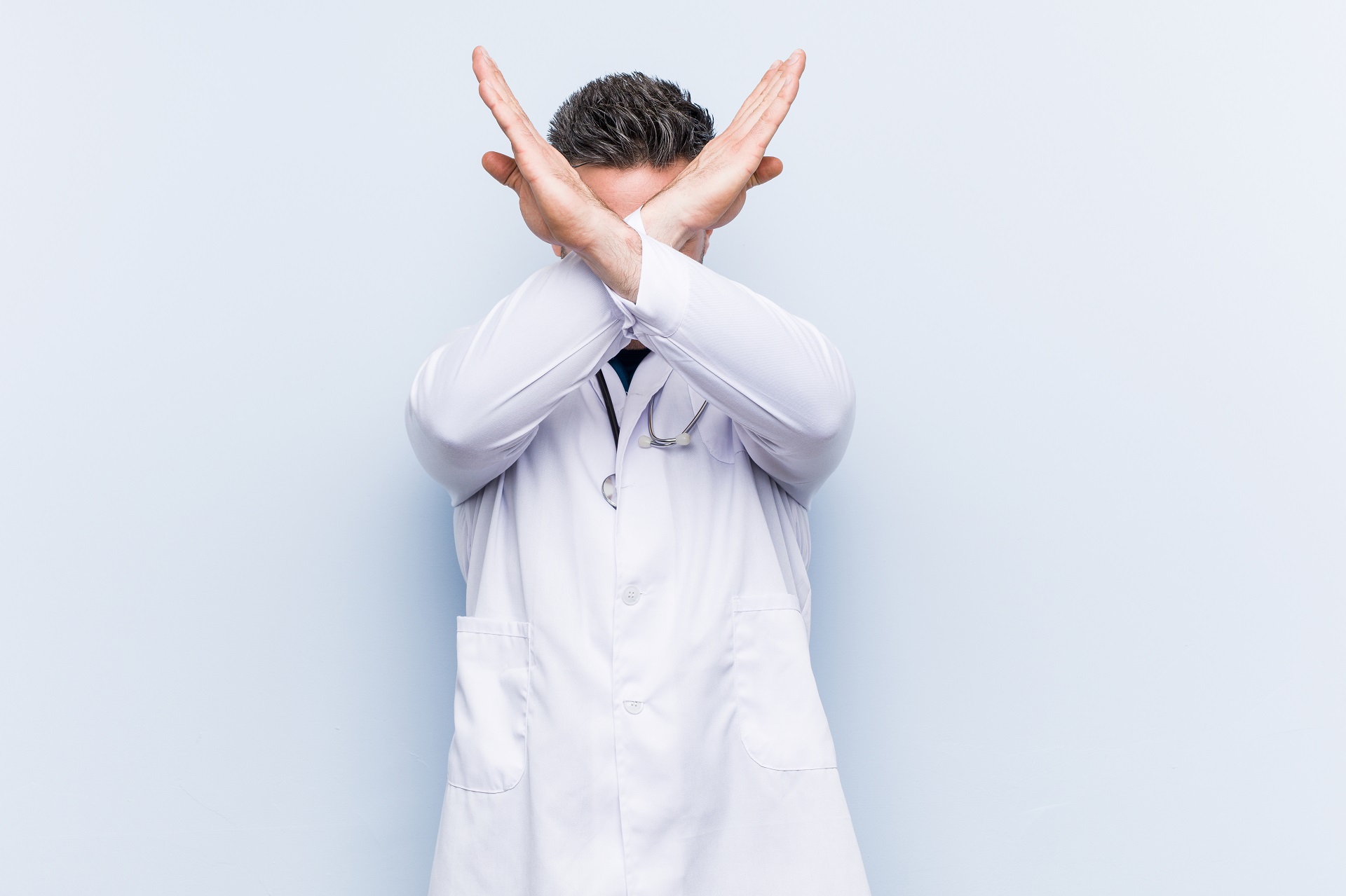 Ein junger Arzt in weißem Kittel versucht sich zu verteidigen indem er seine Arme vor seinem Gesicht verschränkt.