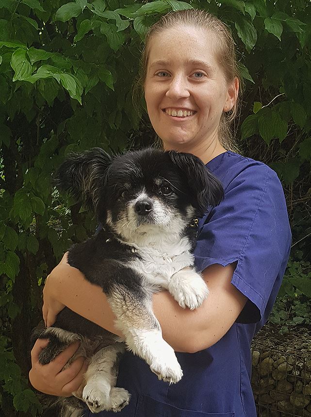 Corinne Leiner trägt einen dunkelblauen Kassak und lächelt in die Kamera. Auf dem Arm hält sie einen kleinen schwarz-weißen Hund.