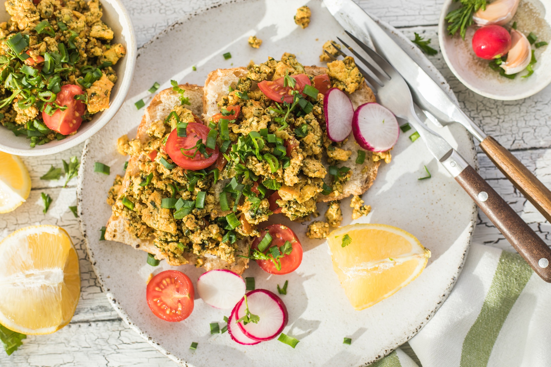 Aufnahme von einem knusprigen Toastbrot mit Rührei aus Tofu und Tomatensalat. Über dem Tofu ist Schnittlauch gestreut. Das Essen ist auf einem Teller angerichtet und daneben liegen ein Set Messer und Gabel.