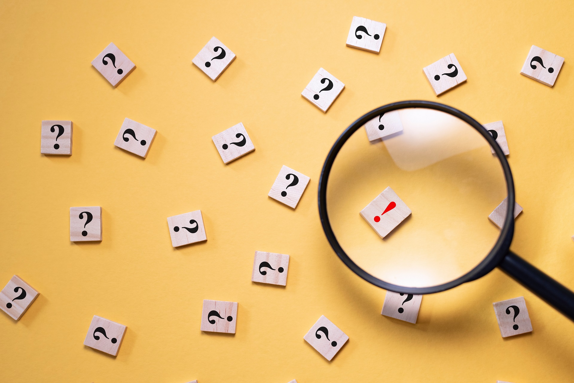Mit einer Lupe wird ein Holzstein mit einem roten Ausrufezeichen vergrößert dargestellt. Um diesen Stein herum liegen nur Steine mit schwarzen Fragezeichen auf gelbem Hintergrund.