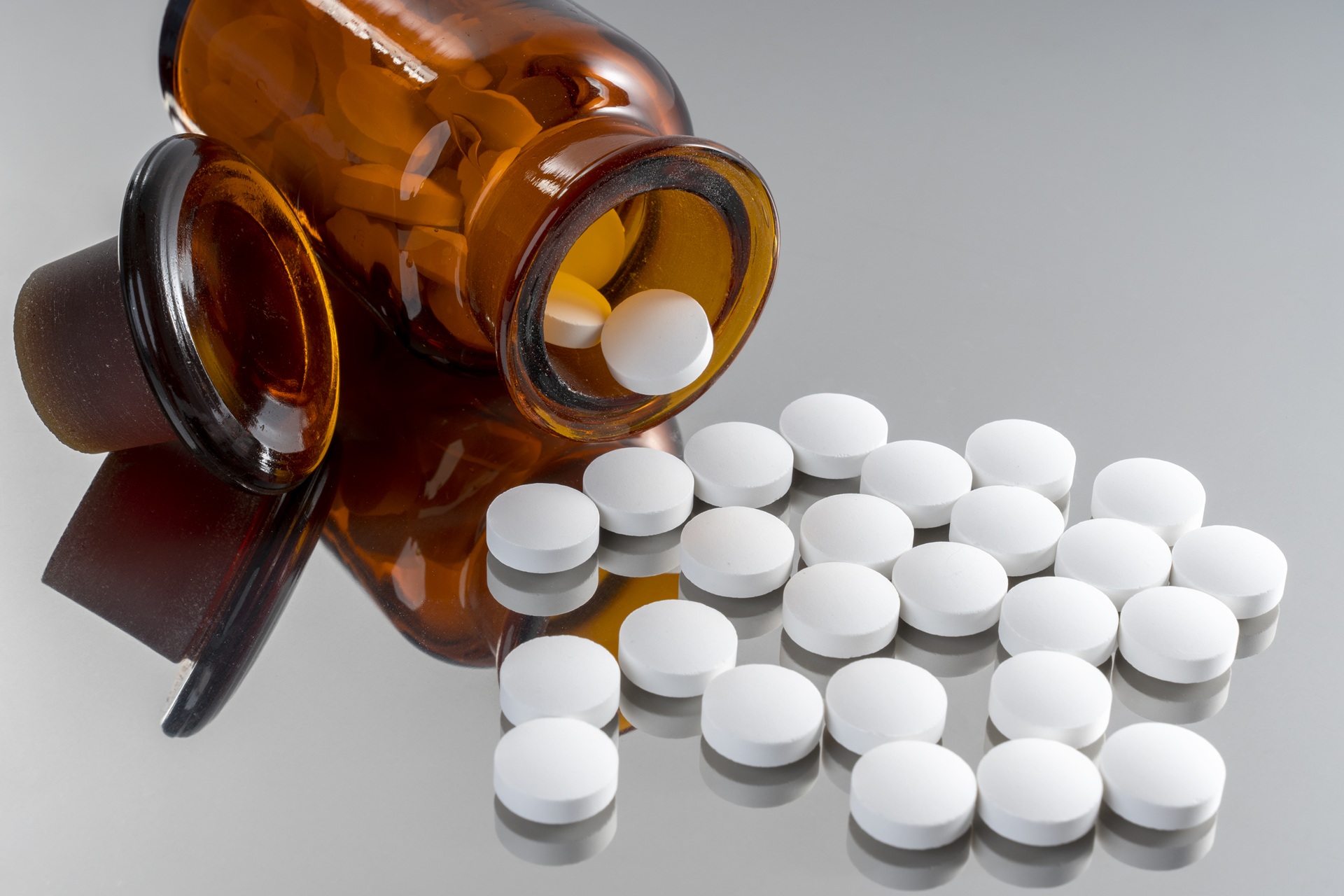 Auf einer silbernen Unterlage liegen weiße Tabletten. Außerdem liegt eine umgekippte Tablettenflasche hinter den Tabletten. In der Flasche befinden sich auch noch weiße Tabletten. Die Flasche ist aus dunklem Glas. 